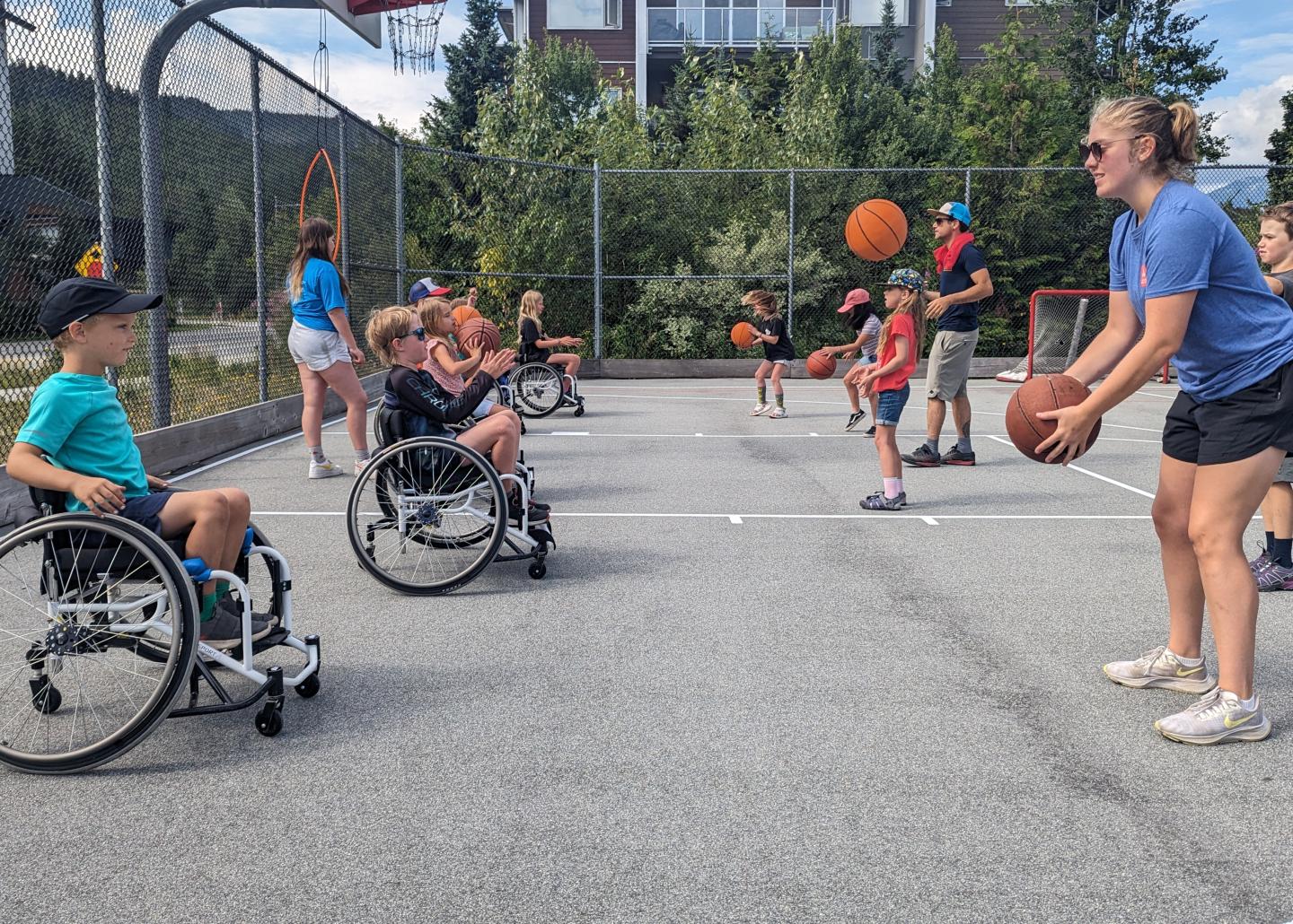 Children in wheelchairs playing basketball on an outdoor court with a smiling coach.